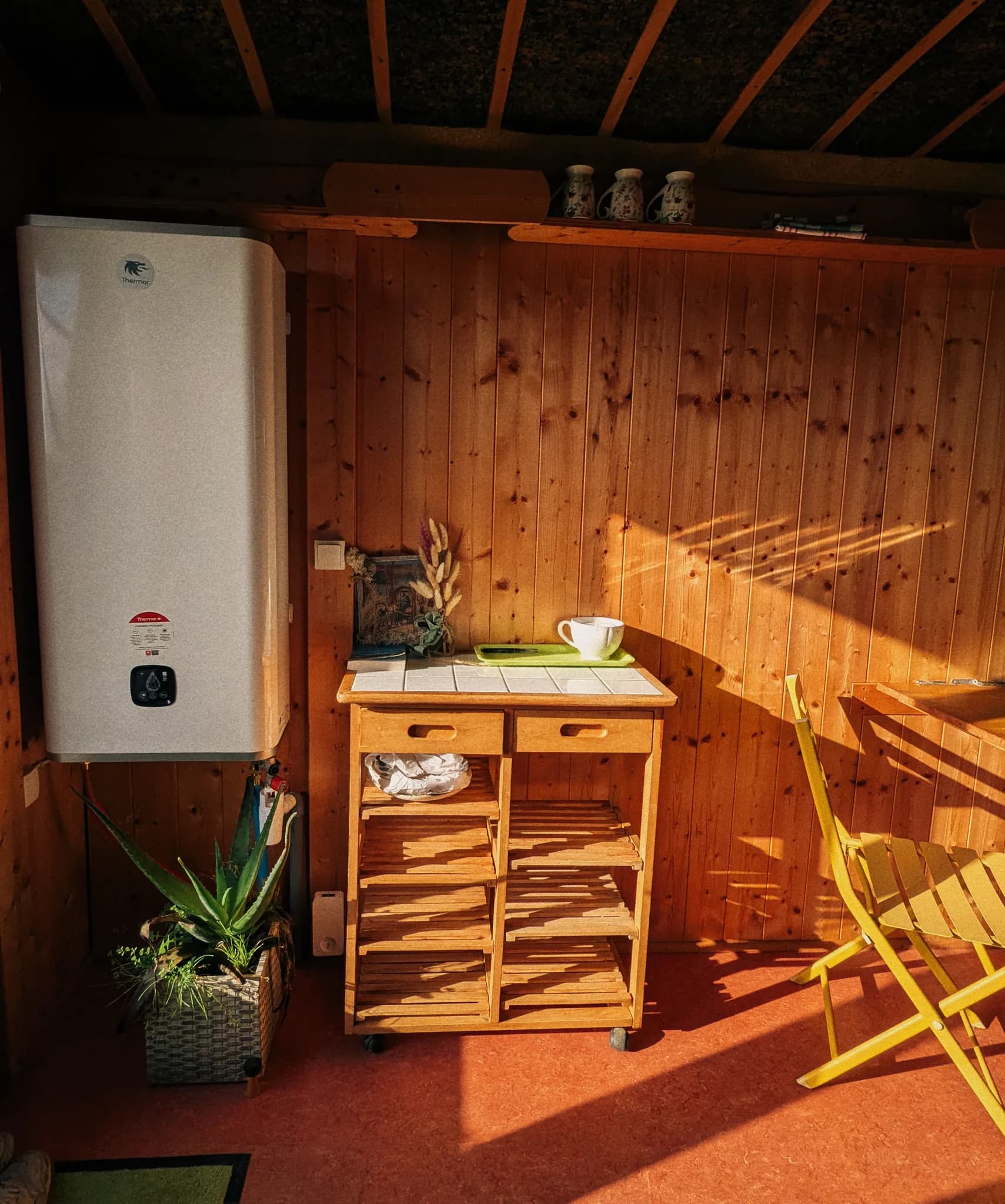 Cozy rustic kitchen with wood paneling, water heater, mobile cart with teacup, yellow chair and warm sunlight streaming in