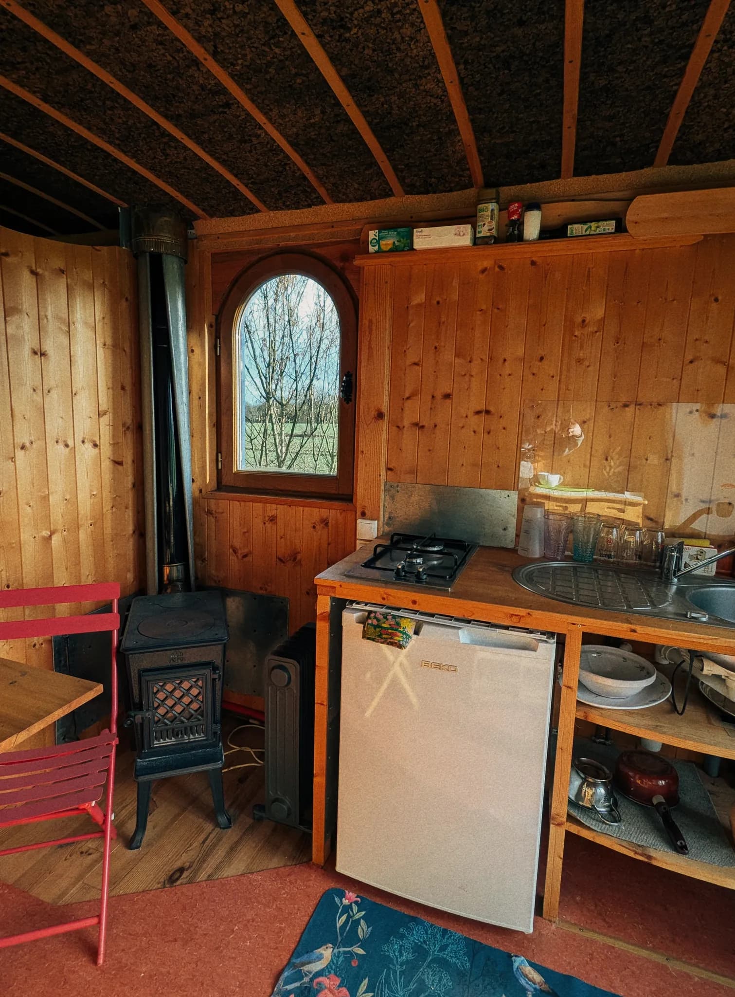 Cozy rustic cabin kitchen with wood-burning stove, arched window, compact appliances and warm wooden interior