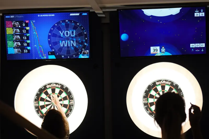 Electronic dart boards with digital displays showing game progress, players throwing darts in modern entertainment venue