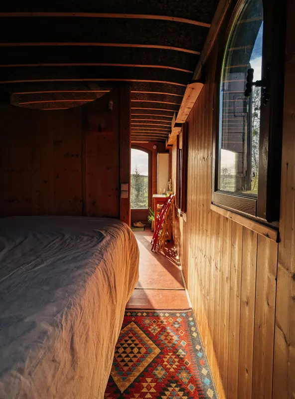 Cozy wooden glamping wagon interior with arched ceiling, colorful kilim rug, bed, and countryside views through arched windows
