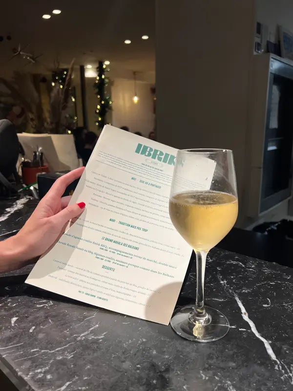 Guest holding Ibrik Kitchen menu with a glass of white wine at a marble bar in a warmly lit restaurant.