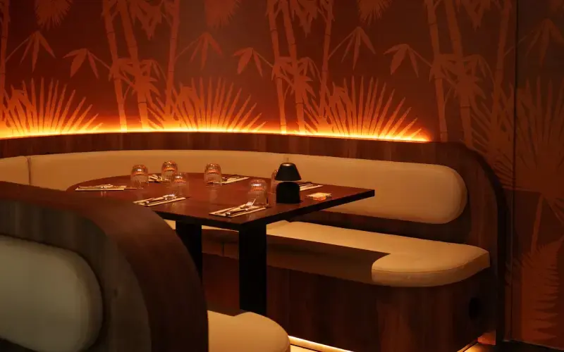 Intimate restaurant booth with cream leather seating, bamboo wall art, warm amber lighting, and elegantly set table for four.