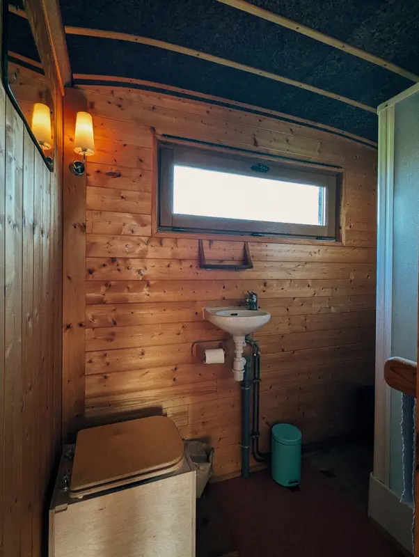 Rustic wooden bathroom interior with wall-mounted sink, toilet, small window, and warm sconce lighting in a cozy cabin setting.