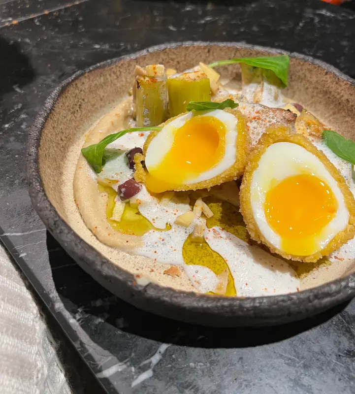 Crispy fried soft-boiled eggs over creamy hummus with olive oil, herbs, and vegetables in a rustic ceramic bowl