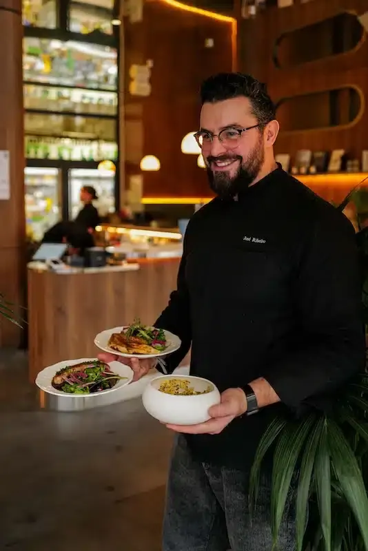 Chef Joel Ribeiro smiling and holding two plated dishes in a warm, modern restaurant dining room.