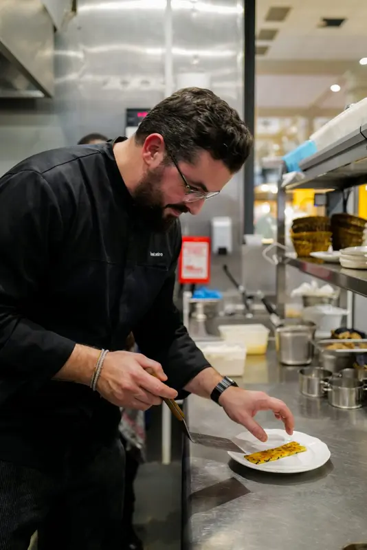 Chef plating a dish in a professional restaurant kitchen, carefully arranging food on a white plate.