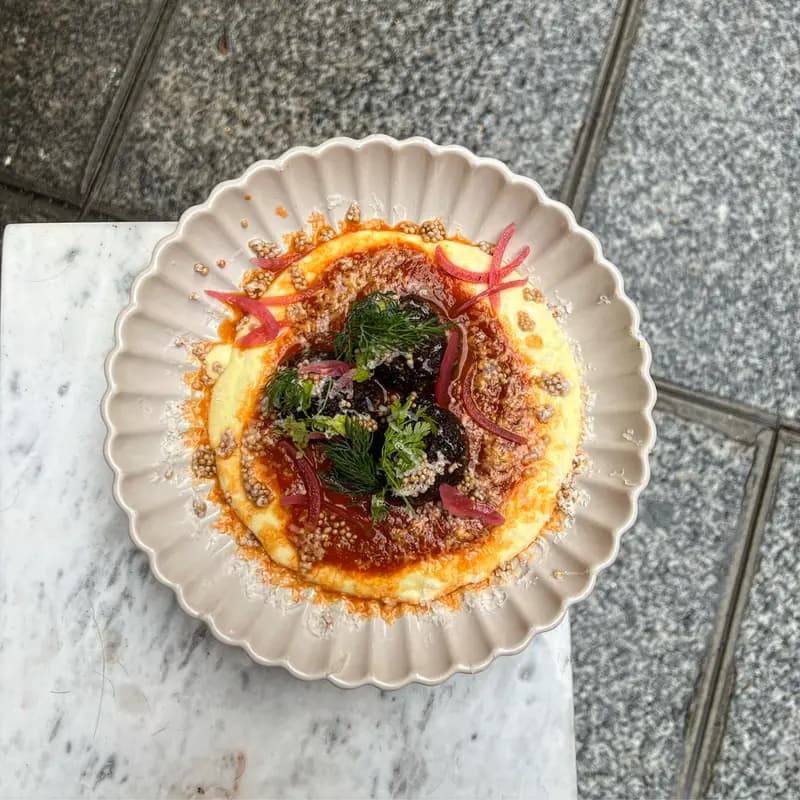 Creamy polenta topped with braised meatballs, tomato sauce, pickled onions, fresh herbs, and grated cheese on a scalloped plate.