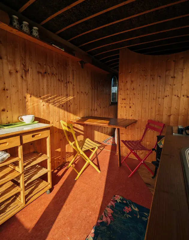 Cozy wooden cabin interior with yellow and red folding chairs, rustic dining table, warm sunlight streaming through walls