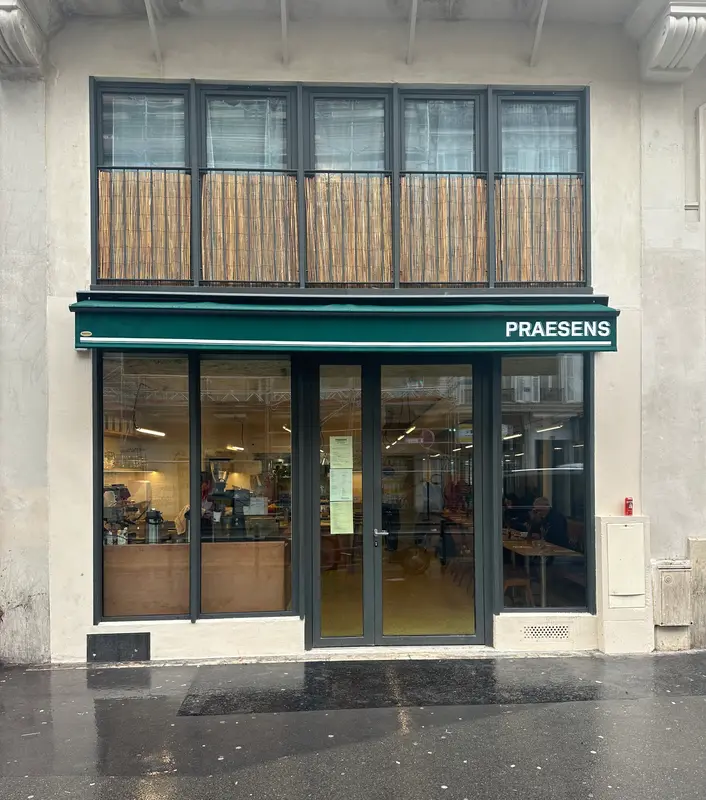 Praesens restaurant storefront with green awning, large windows, and cozy interior seating visible through glass facade.